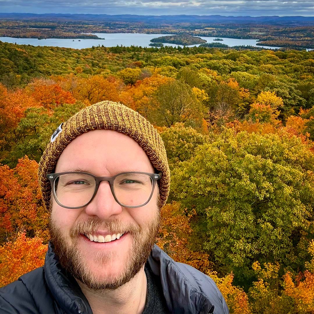 Carson smiles and slightly squints at the camera, wearing a yellow touque, blue windbreaker, and glasses. A wide view of autumn leaves fills the landscape behind; vivid orange, yellow, red, and green leaves stretch out to a view of Lac Blue Sea in Gatineau, Quebec.